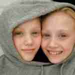 Charming close-up of smiling twin girls with blonde hair sharing a grey hoodie indoors.